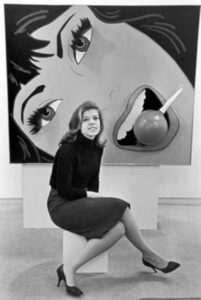 Photographic portrait of Marjorie Strider sitting in front of her painting Girl with Radish (1963).